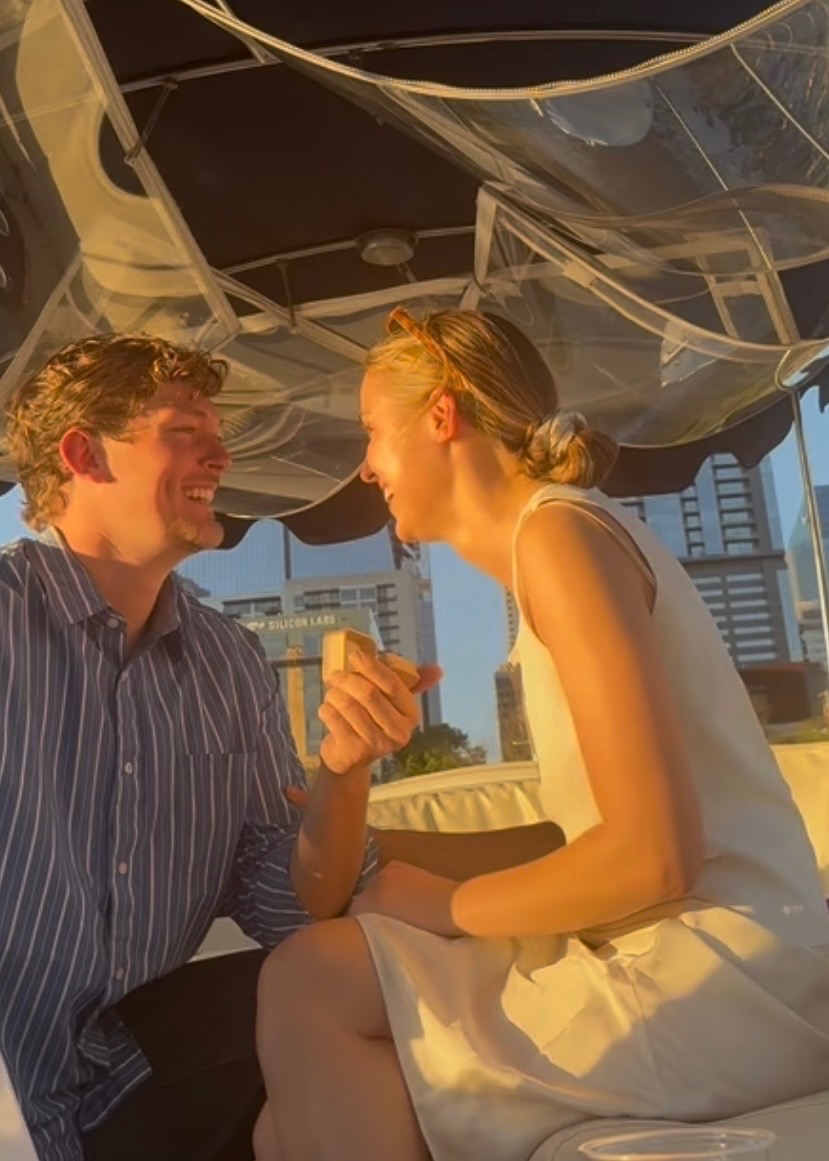 Kai holding out a ring box to Madeleine, laughing in a white dress, on a boat at golden hour with the Austin skyline behind them