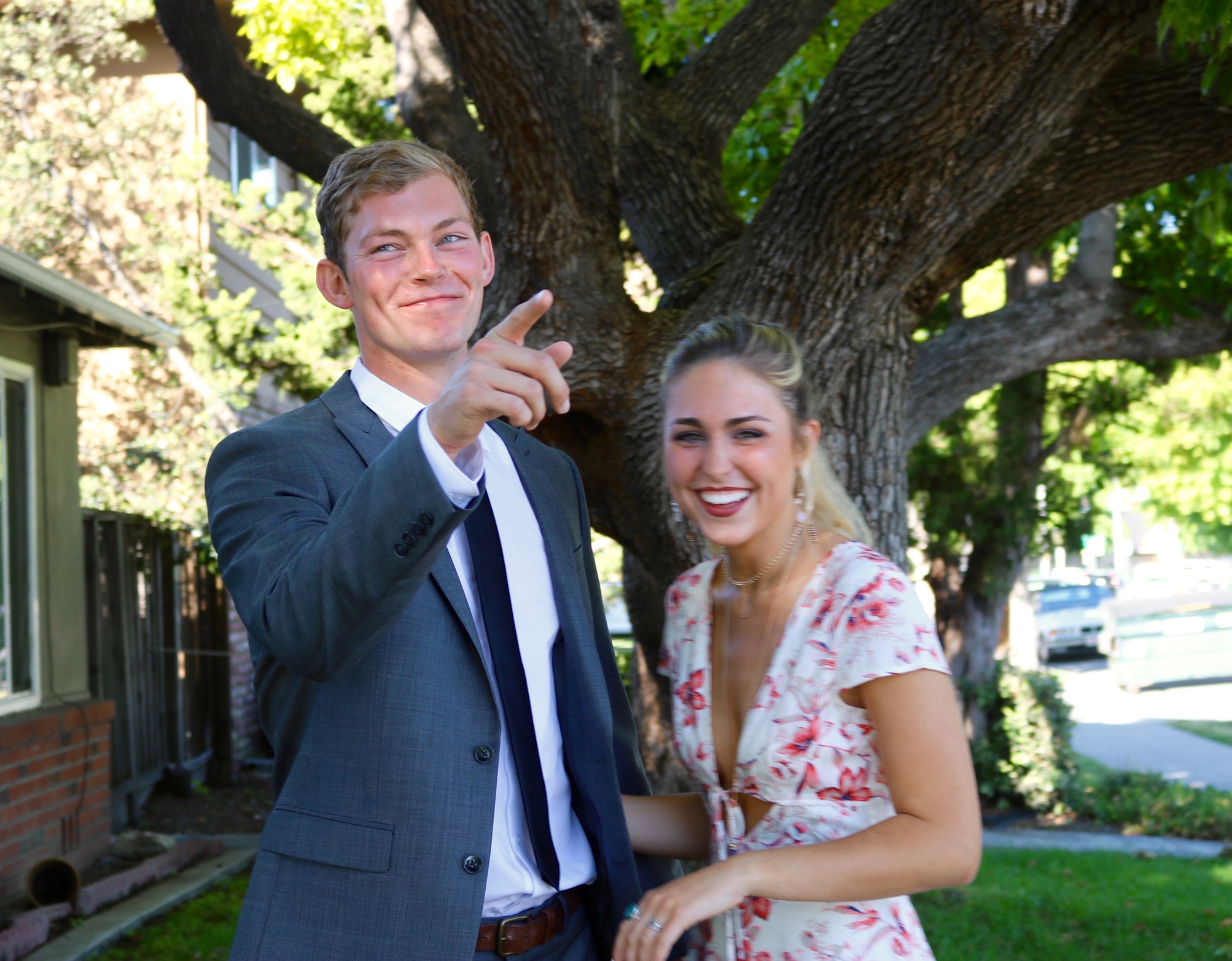 Kai in a gray suit pointing and smiling beside Madeleine, laughing in a floral dress, under a big oak tree
