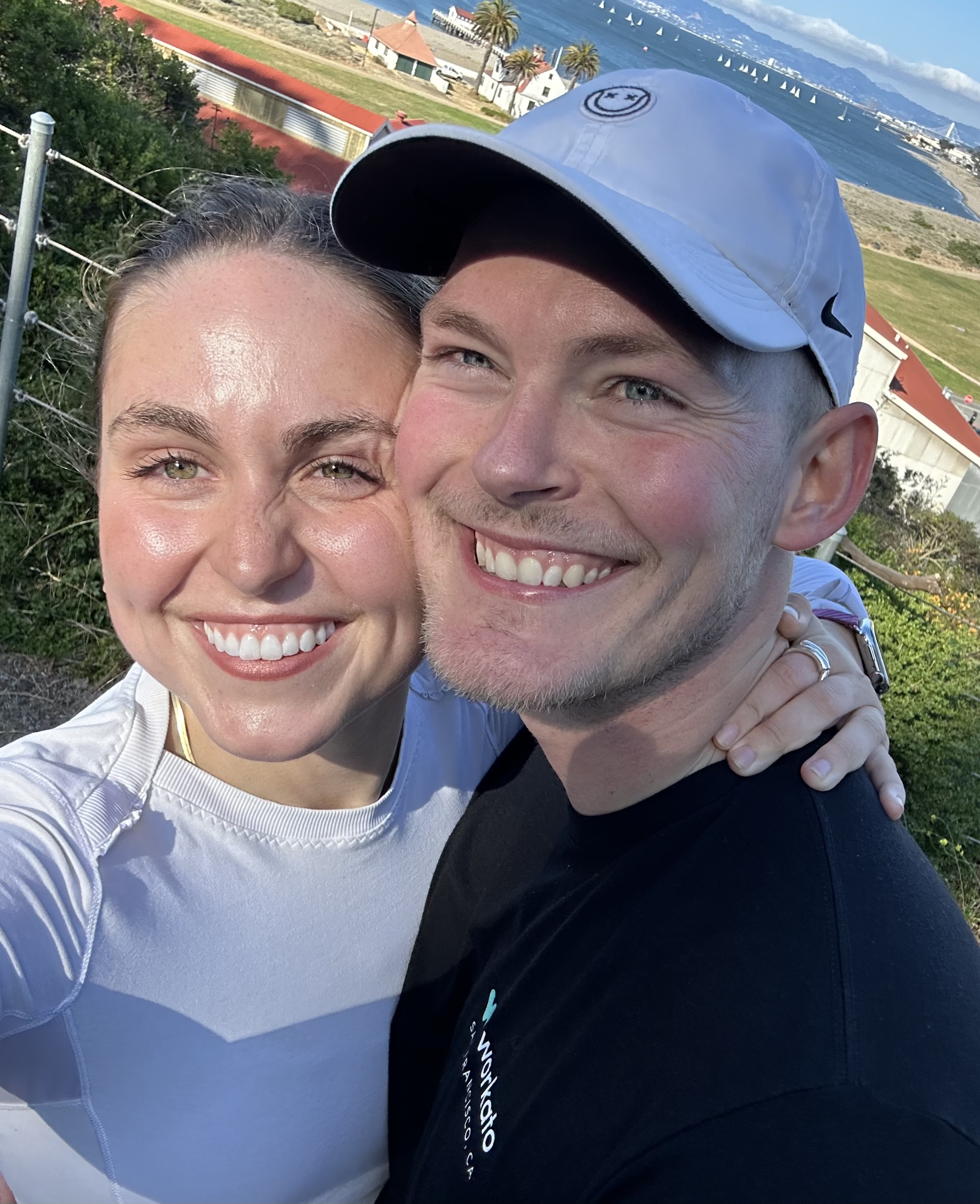 Madeleine and Kai smiling in a selfie on a hike above the San Francisco Bay, with sailboats on the water behind them